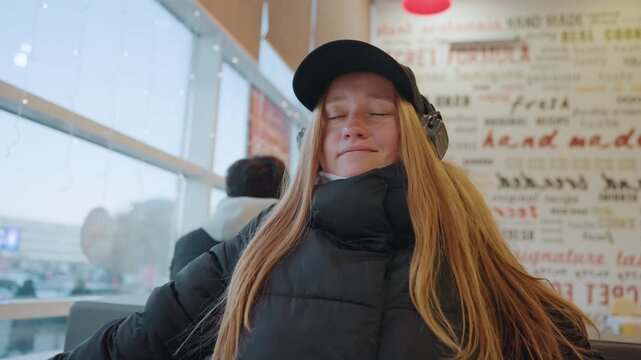 White woman closeup at airport window, introspective passenger gazing toward tarmac with headphones and cap, soft light on freckled skin, contemplative serene mood during travel layover.