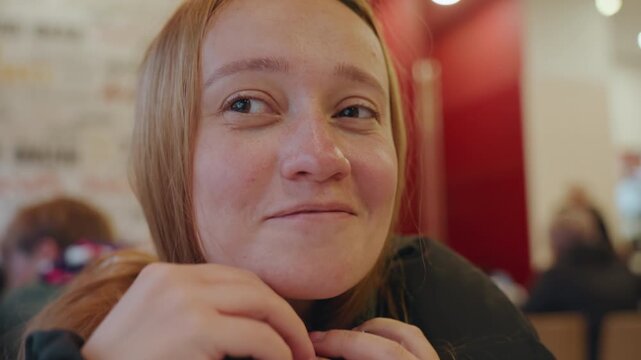 White woman in busy cafe student adjusts headphones while scanning crowd, tense expression, red interior backdrop, candid closeup portrait conveying vigilance and concern about snatcher risk