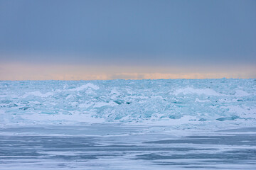 Obraz premium A breathtaking winter landscape featuring vast piles of translucent blue ice shards pushed up onto the shoreline of a frozen Great Lake. In the distance, a soft band of peach-colored light glows.
