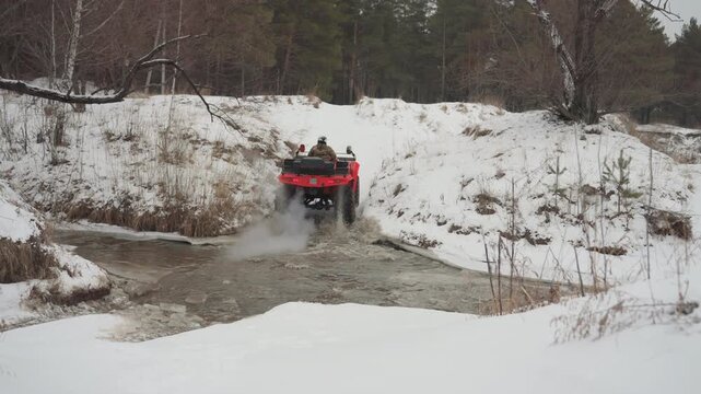 Red atv splashing through frozen creek, rider charges down snowy bank into shallow water, spray and mud fling, pine forest backdrop, overcast sky, fast offroad motion, dynamic wake and dramatic winter