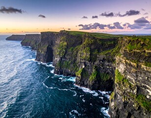 Obraz premium Aerial view of high green cliffs meeting vast blue ocean under a colorful sunset