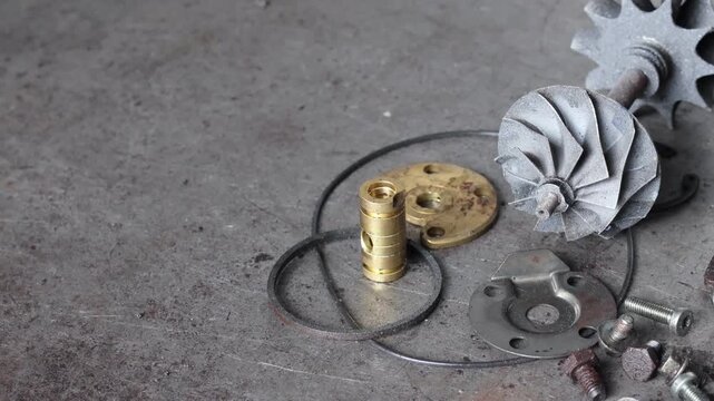 Panning shot of disassembled turbocharger parts including housing, turbine wheel, and brass bearings arranged on a rustic metal workshop floor.