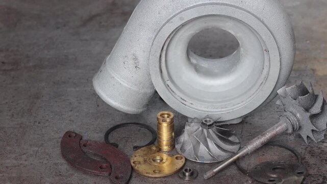 Panning shot of disassembled turbocharger parts including housing, turbine wheel, and brass bearings arranged on a rustic metal workshop floor.