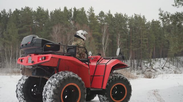 Red atv rider in snowy forest, helmeted driver in camouflage jacket seated on powerful red quad with oversized tires, rear cargo box on rack, slow patrol along pinelined trail, fresh tracks in soft