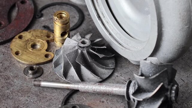 Panning shot of disassembled turbocharger parts including housing, turbine wheel, and brass bearings arranged on a rustic metal workshop floor.