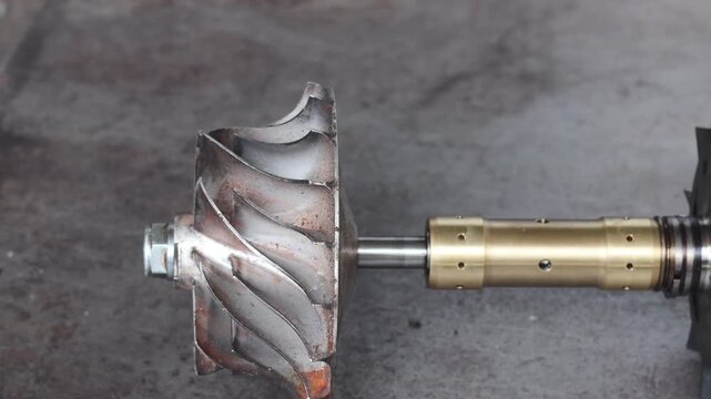 Panning shot of a complete turbocharger shaft assembly with turbine and compressor wheels and brass bearing journal lying on a rustic industrial workshop floor.