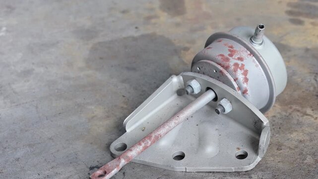 Panning shot of a rustic mechanical turbocharger wastegate actuator assembly with a rusty rod and mounting bracket on a weathered workshop surface.