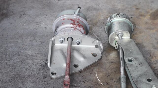 Panning shot of two metallic turbocharger wastegate actuators on a rustic workshop surface, showing one weathered unit with rust and one clean unit with its mounting bracket.