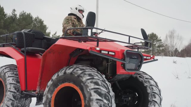 Red atv in snowy field rider in camouflage jacket and helmet navigates icy trail, close up of chunky tires throwing snow, exhaust steam and undercarriage detail, rear cargo rack and forest backdrop,