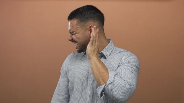 Man grimacing with hand pressed against ear and clenched jaw in studio with terracotta backdrop; discomfort.
