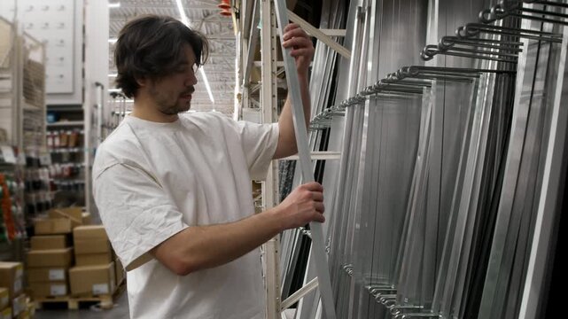 Man reaches up for long aluminum profile from rack in hardware store. He grips silver beam amid glass sheets and shelves.