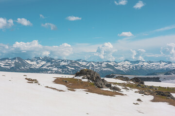Scenic alpine landscape with sunlit snowy field on stony hill with view to big mountain range with forest and snow far away under cloudy sky. Stone outcrops among snows in sunlight in high mountains. © Daniil