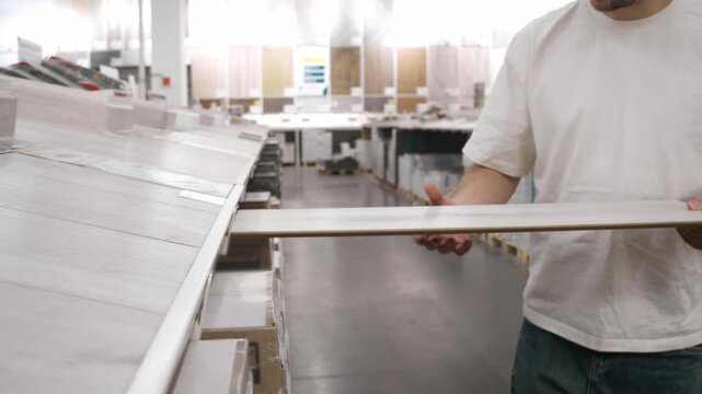 Wide shot shows man standing at laminate rack in hardware store, lifting long flooring plank to inspect grain pattern and thickness before purchase decision.