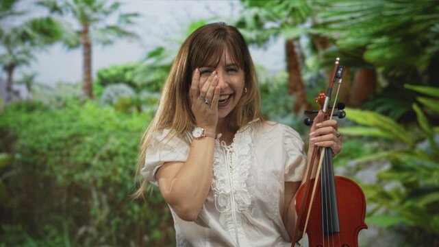 Woman laughing with hand covering face, holding violin and bow in forest among green foliage and palm trunks; joy.