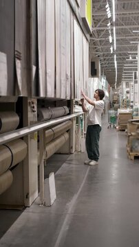 Shopper evaluates vinyl flooring sample in construction market rack. Fingers feel material near stacked rolls. vertical