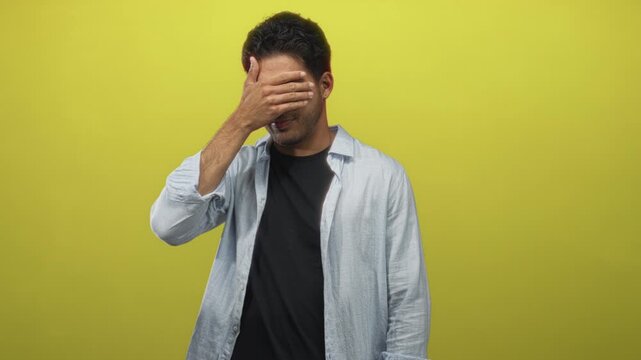 Man covering face with hand in studio against bright yellow backdrop, wearing light shirt over black tshirt, standing and looking down; embarrassment.