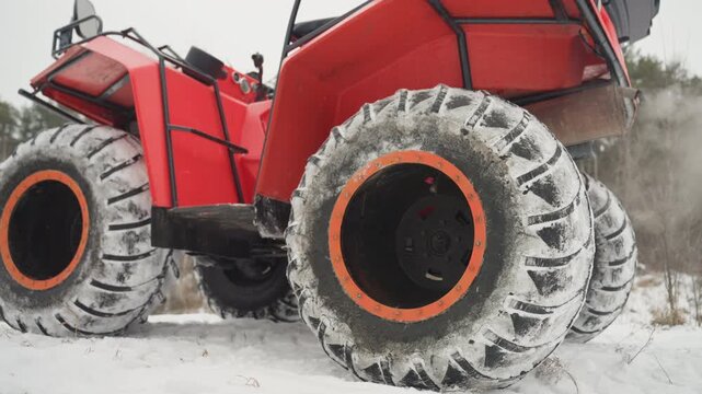 Red ATV wheels kicking up snow, low angle closeup of oversized studded tires covered in frost with camouflageclad rider beside vehicle, winter forest backdrop, engine steam, rugged offroad terrain,