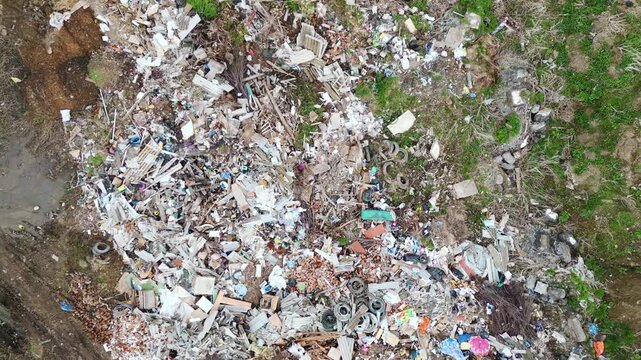 Aerial view shows scattered trash in a grassy area. Waste piles mix plastic, paper, and broken items. Muddy ground edges the chaotic trash pile. No people appear in the scene