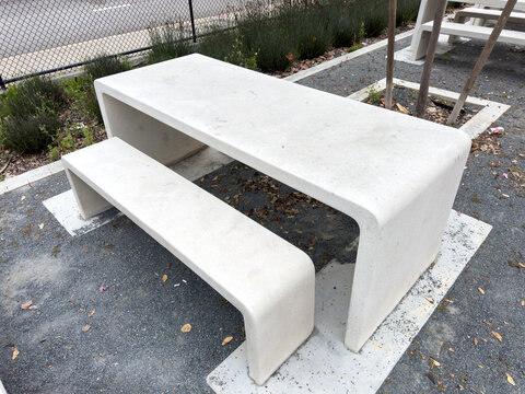 Smooth concrete benches and tables sit outdoors on gravel and paved squares the minimalist design features curved edges and a modern appearance with plants in the background