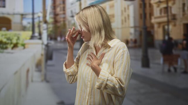 Young woman pinches nose with hand pressed to chest on a sunlit street among buildings and lampposts; discomfort concern.