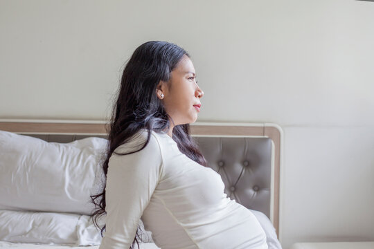 Pregnant Asian Woman Sitting on Bed with a Pensive and Thoughtful Expression
