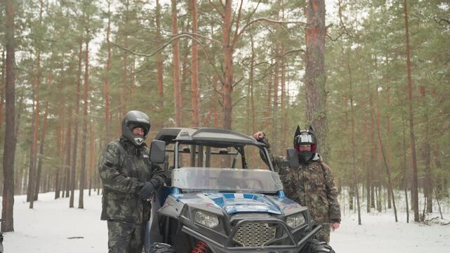 Two riders beside blue atv in snowy pine forest, helmeted drivers stand next to utility terrain vehicle and snowmobile, evergreen trunks and fresh snow create rugged atmosphere, visible tire tracks
