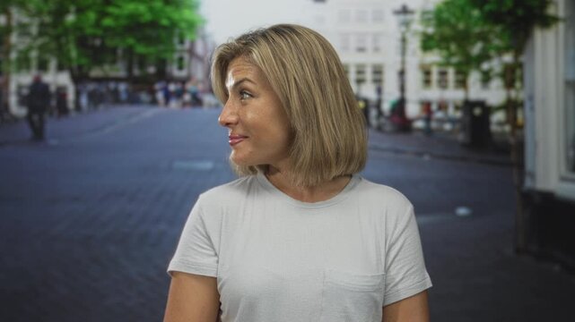 Woman points thumb to the side on a busy city street, standing outdoors in a white t shirt and bob haircut while looking to her left; confidence.