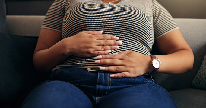 Hands, stomach ache and woman in home with pain, endometriosis or menstrual cramps on sofa. Living room, sore abdomen and sick person with constipation, food poisoning and bloated with gas in house