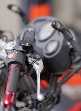 Close-up of a motorcycle handlebar shows the right grip, brake lever, fluid reservoir, and kill switch, with analog gauges slightly blurred in the background, 2026