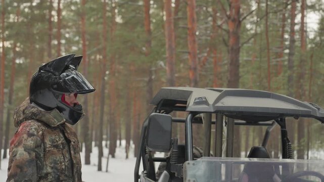 Snowy forest atv rider with cat ear helmet steadying utv door, camo jacket and red balaclava, light snow on pine trees, playful adventurous pause before offroad run, hand on mirror and close helmet