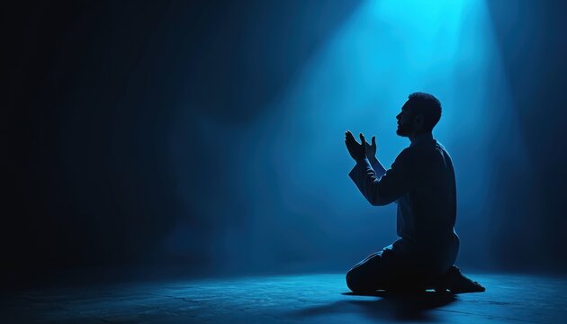 Man kneels in silhouette praying with hands raised. Blue light shines from above. Religious devotion and faith concept. Muslim man praying in dark.