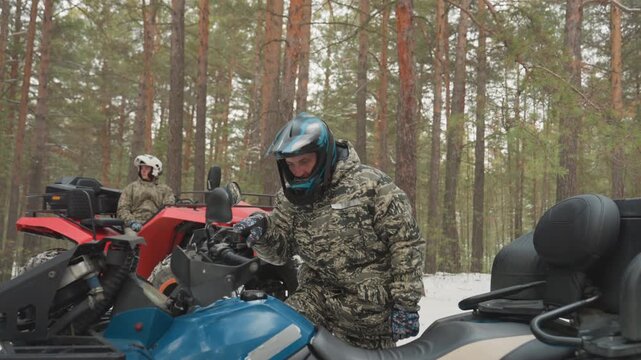 Snowy pine forest duo prepping atvs, helmeted rider inspecting machine while companion waits, red and blue quads parked on trail, hands checking throttle and mirror, teamwork before launch, crisp cold