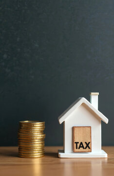 Small house model with TAX sign sits next to stack of gold coins. Conceptual image for property taxes mortgage costs home investment expenses and wealth.