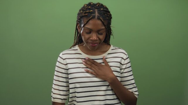 Woman with braided hair in striped shirt placing hand on chest in green studio; quiet gratitude reflection.