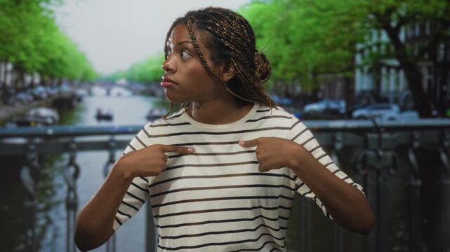 African american woman in striped shirt points fingers to chest and looks wide eyed on street beside an amsterdam canal bridge; surprise.