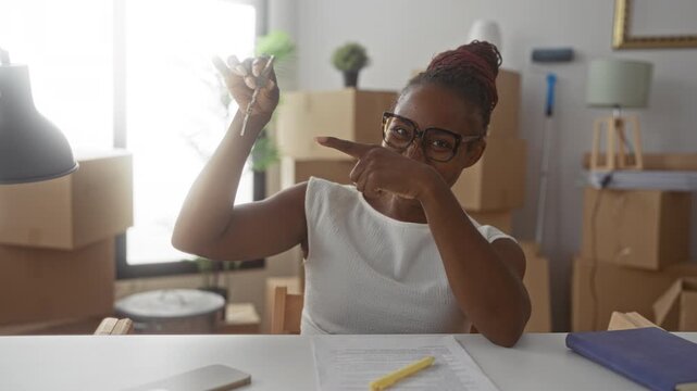 Young woman holding house keys, points finger at key over paperwork and moving boxes inside a building; joy new beginning.