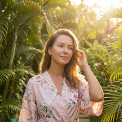 Beautiful relaxed woman wearing pink floral robe standing outside feeling peaceful during vacation travel enjoying morning sunlight surrounded by green tropical palm leaf garden setting nature