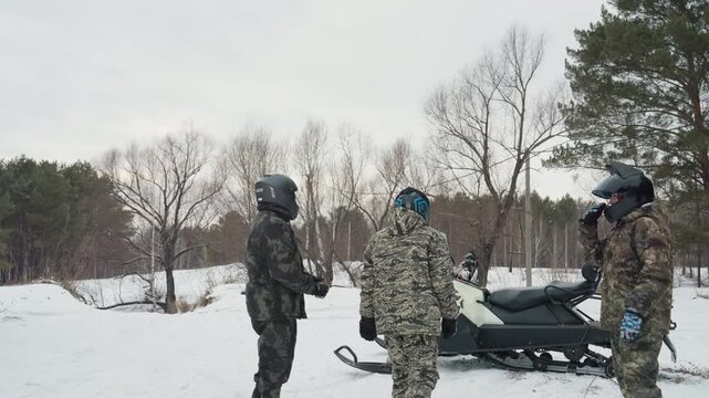 White riders by snowmobile and ATV in snowy forest, three camouflaged riders in helmets converse beside parked snowmobile and distant ATV, guide assigning trail roles, visible breath in cold air, pine