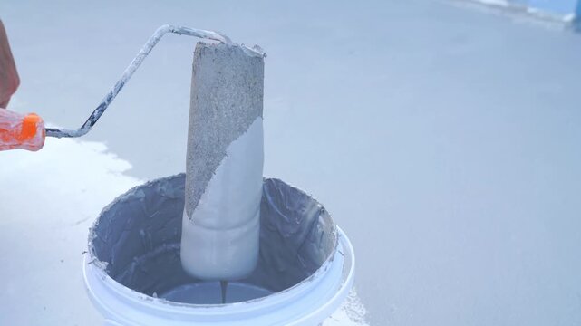 A close-up of a hand dipping a paint roller into a bucket of grey waterproof coating. Applying a protective layer to a concrete floor or rooftop. Industrial construction and home repair concept.
