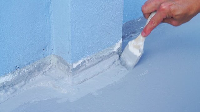 Close up of a hand applying grey waterproofing coating to a blue wall corner using a brush. House renovation and roof leak repair maintenance concept. Professional construction work in progress.
