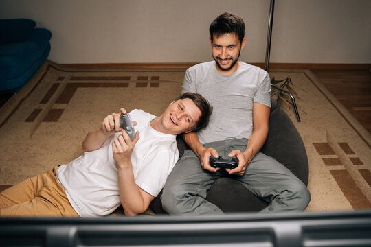 Two gay men relaxing at home, enjoying casual evening together, sharing comfortable moment while playing together video games with console controllers. Concept of modern online entertainment.