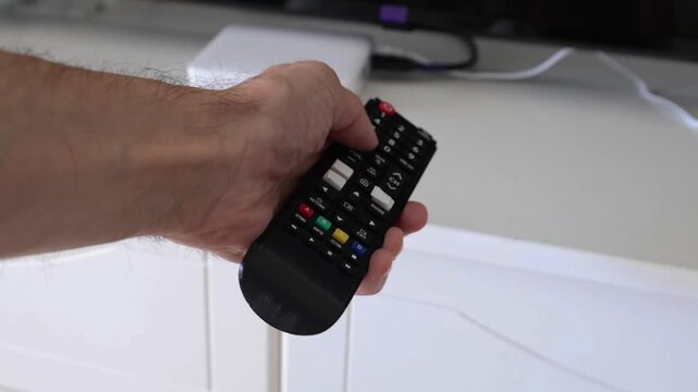Male hand holding a TV remote control and pressing different buttons with thumb. Media box and television visible in the background. Concept of controlling TV and home entertainment.