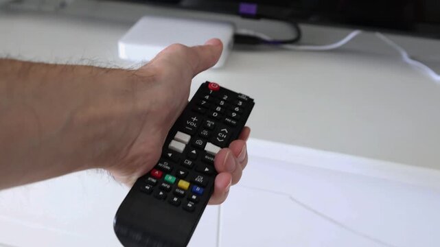 Male hand enters the frame holding a TV remote control, points it toward the television, and presses the red power button. Media box and TV visible in the background. Concept of turning on TV and home