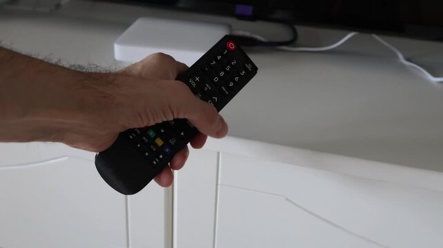 Male hand holding a TV remote control and pressing the volume up button several times. Media box and television visible in the background. Concept of adjusting sound and home entertainment.