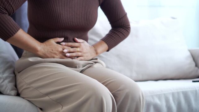 Close-up of a woman sitting on a sofa clutching her stomach in pain, suffering from abdominal cramps, menstruation, or digestive issues at home. Concept of female health and stomachache