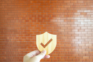 Hand holding a wooden shield with checkmark sign against a brown brick wall. Conceptual image for...