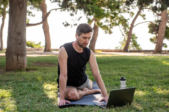 Man practicing yoga outdoors on mat with laptop in park