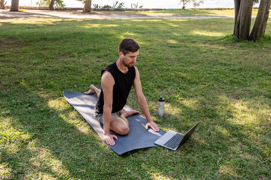 Man practicing yoga outdoors on mat with laptop in park