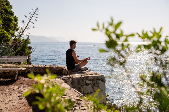 Man meditating in yoga pose on rocky ledge by the sea