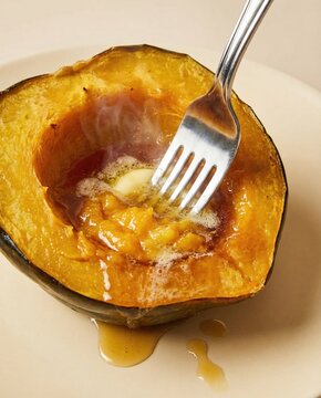 Steaming half of roasted acorn squash with melting butter and syrup, pierced by a fork on a light beige plate